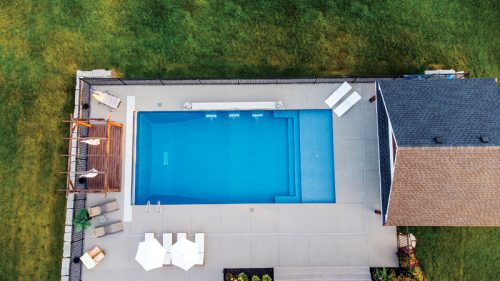 An aerial view showcases a residential backyard featuring a blue swimming pool surrounded by a concrete patio. There are several lounge chairs positioned on one side, along with two white umbrellas. A brown wooden deck is visible near the pool, and a house with a gray roof is situated to the right, complemented by greenery in the surrounding area.