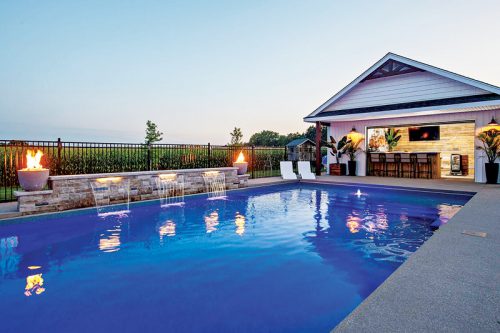 A beautiful outdoor scene showcasing a rectangular blue swimming pool surrounded by a smooth patio. To the left, a stone water feature flows into the pool, and two flames flicker from decorative pots. A modern pavilion with a bar area and stylish seating is situated in the background, complemented by lush greenery, creating a welcoming atmosphere.