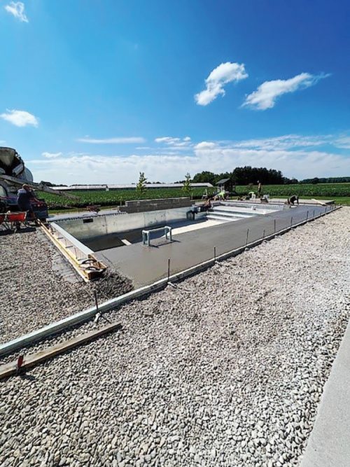 A construction site features freshly poured concrete, forming a pool or basin. Workers are present, actively engaging in tasks around the site. In the background, fields and trees are visible under a clear blue sky, with fluffy white clouds scattered above.