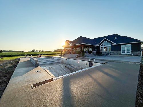 A picturesque scene featuring a home in a rural landscape during sunset. The house has a dark roof and large windows, providing a welcoming appearance. In the foreground, a pool is under construction with cement borders, while the sun casts a warm glow over the property. Lush green fields stretch in the background.