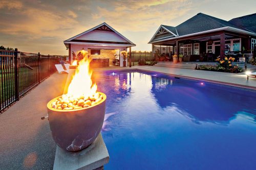 A vibrant sunset casts an orange glow over a tranquil backyard scene featuring a blue swimming pool. In the foreground, a modern fire feature with flames rises from a round stone basin filled with pebbles. The pool is bordered by a black fence, and a well-designed house with large windows is visible in the background, complemented by landscaped flower beds and leisure chairs.