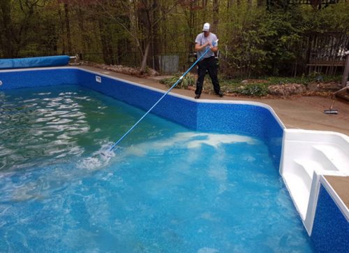 Person cleaning a residential swimming pool using a long pole with a net, surrounded by trees and a paved pool deck.