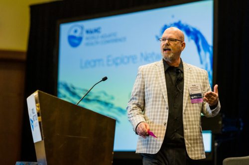 Speaker presenting at the Water Quality Association Health and Sustainability Conference, standing in front of a podium and a screen with the text 'Learn. Explore. Network.' in the background.