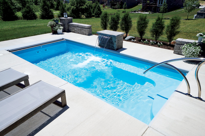 A sparkling blue rectangular pool is surrounded by light-colored stone decking. Two lounge chairs are positioned to the left, while a stone water feature spills into the pool.