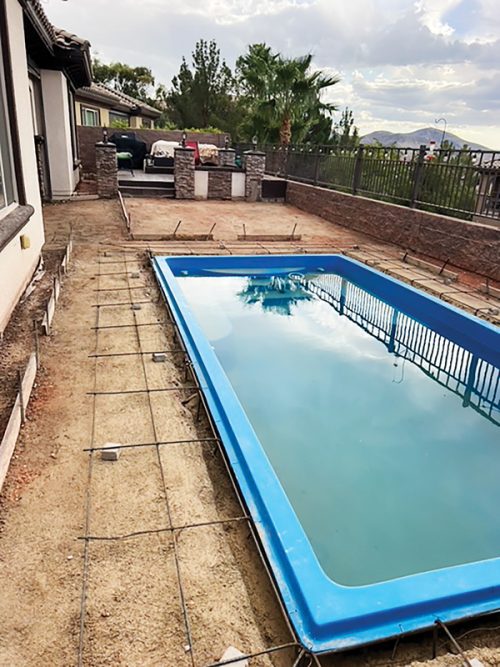 Swimming pool under construction, surrounded by gravel, with a fenced backyard and palm trees in the background.