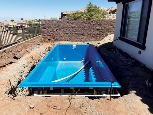 Blue swimming pool partly installed in a backyard with a fence and residential houses in the background.