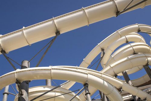 A series of white, winding water slide tubes, partly stacked and interconnected, contrasting against a bright blue sky. The tubes are supported by dark metal structures, showcasing a playful and adventurous water park design.