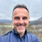 John Bereza stands outdoors in a blue jacket, against a backdrop of mountains and cloudy skies, capturing a moment in nature.