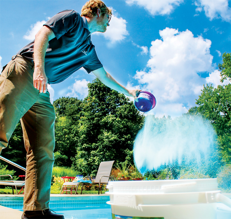 A man in a blue shirt pours blue powder into a pool from a container, surrounded by lush greenery and a bright blue sky.