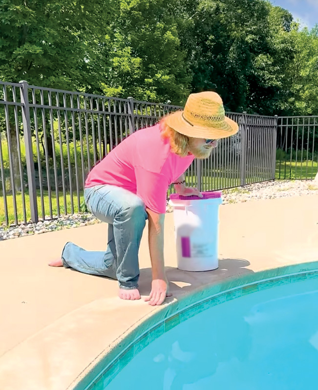 Person in a straw hat and pink shirt kneeling by a swimming pool, with a white bucket that has a purple lid nearby.