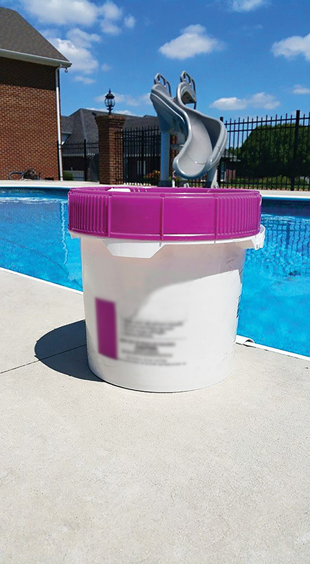 White bucket with a purple lid placed on a poolside deck, with a swimming pool and slide in the background.