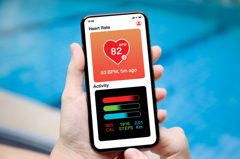 Person holding a smartphone displaying a heart rate and activity tracking app, with a swimming pool in the background.