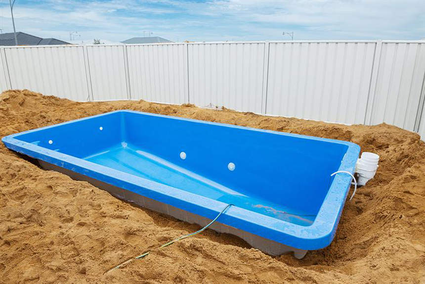 A partially installed blue fiberglass swimming pool shell set in a sandy excavation site, with white PVC plumbing connections visible.