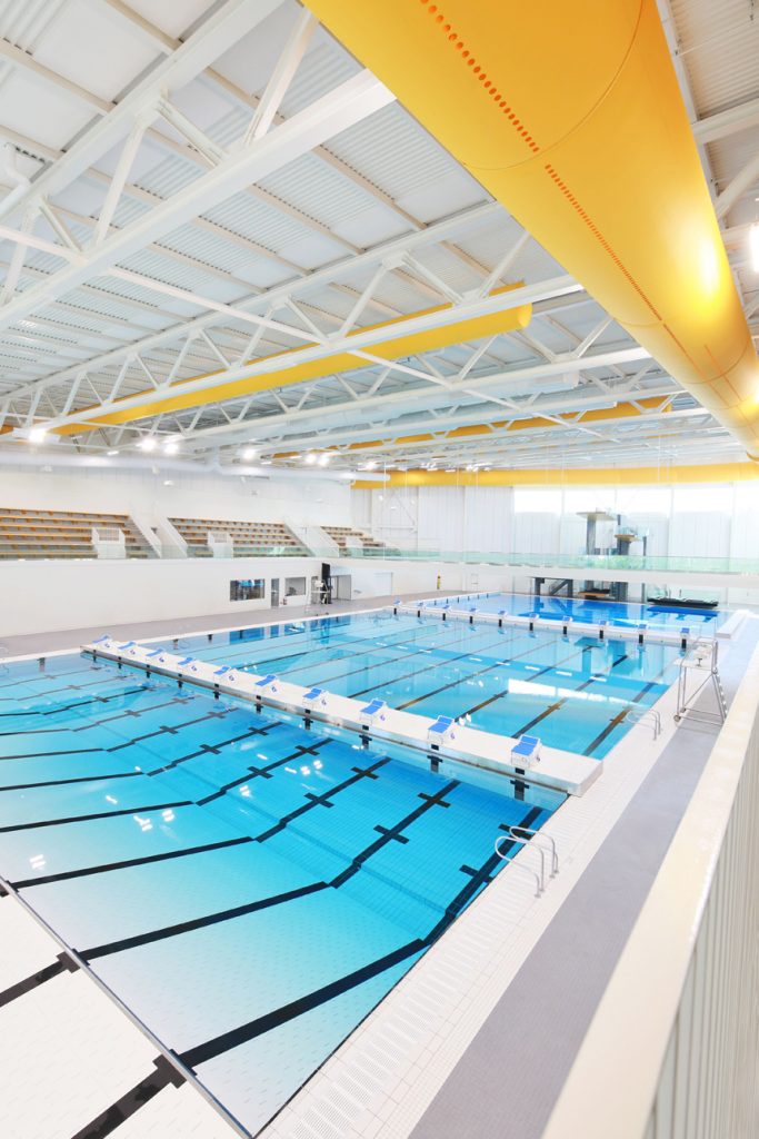 Bright indoor swimming pool with multiple lanes, diving boards, and seating area overlooking the water.