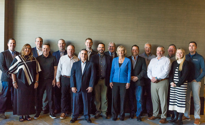 A diverse group of professionals poses together, dressed in business attire, in a well-lit indoor setting against a textured wall.